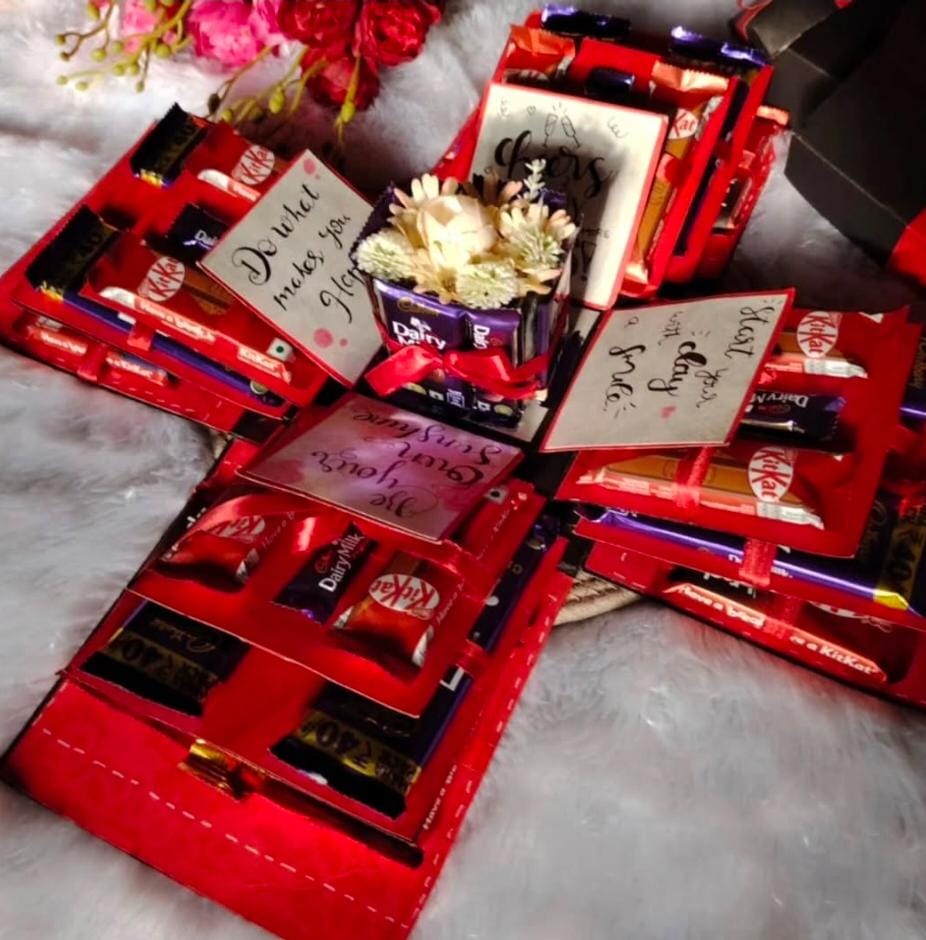 A handmade red explosion box gift filled with chocolates like Dairy Milk and KitKat, decorated with red ribbons, message cards, and artificial flowers in the center. The box opens outward to reveal multiple layers of chocolates and heartfelt notes, creating a beautiful customised hamper made by Aabis Crafty.