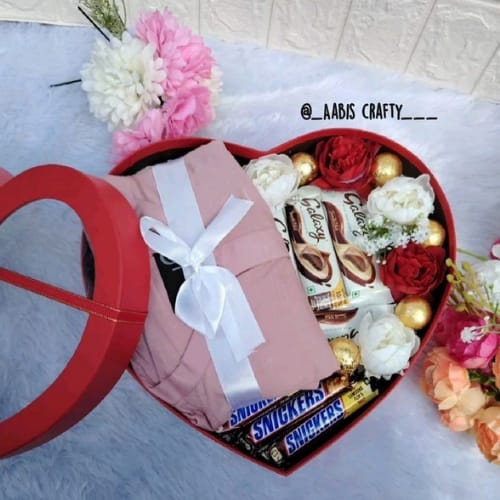 A heart-shaped red gift box filled with chocolates, including Snickers and Galaxy bars, along with white and red artificial roses, golden decorative balls, and a neatly folded pink outfit tied with a white ribbon. The box lid rests beside it, and the tag reads “@Aabis Crafty__,” showcasing a beautifully customised hamper for gifting.